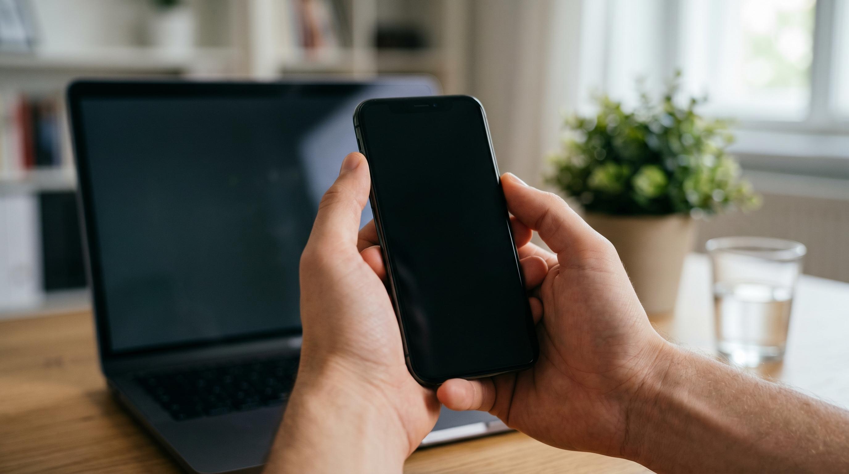 Hands holding a smartphone near a laptop at a desk while linking a device